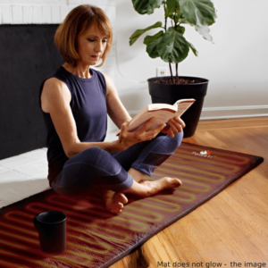 A woman sitting on a ToastiMat Heated Yoga Floor Mat reading a book with a cup beside her.