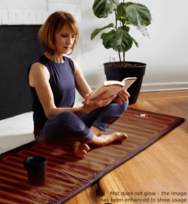 A woman sitting on a ToastiMat Heated Yoga Floor Mat reading a book with a cup beside her.