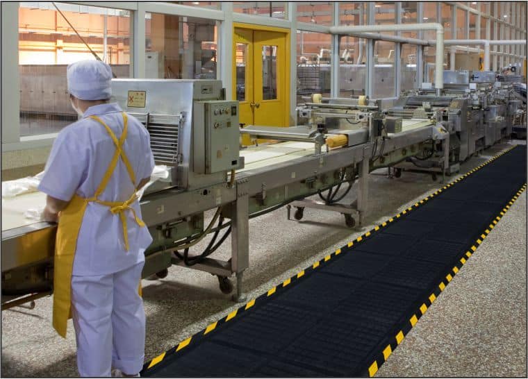 A worker is standing on a Hog Heaven III Linkable Comfort Floor Mat in a factory.