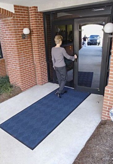 A woman walking into a building with a WaterHog Eco Premier Floor Mat.