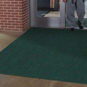 A person standing on a large green WaterHog Squares Fashion mat inside a building with a glass door and brick wall visible.