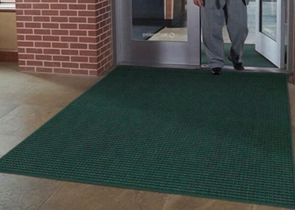 A person standing on a large green WaterHog Squares Fashion mat inside a building with a glass door and brick wall visible.